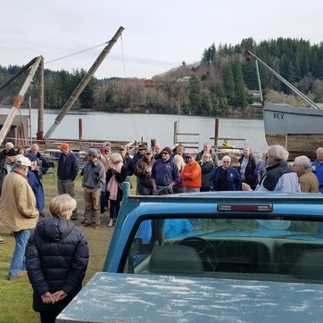 A group of people gathered outdoors near water with a crane and trees in the background.