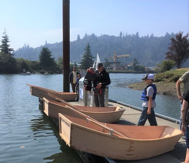 People by the water with wooden boats, likely preparing for boating or a boat launch, in a serene, tree-lined area.