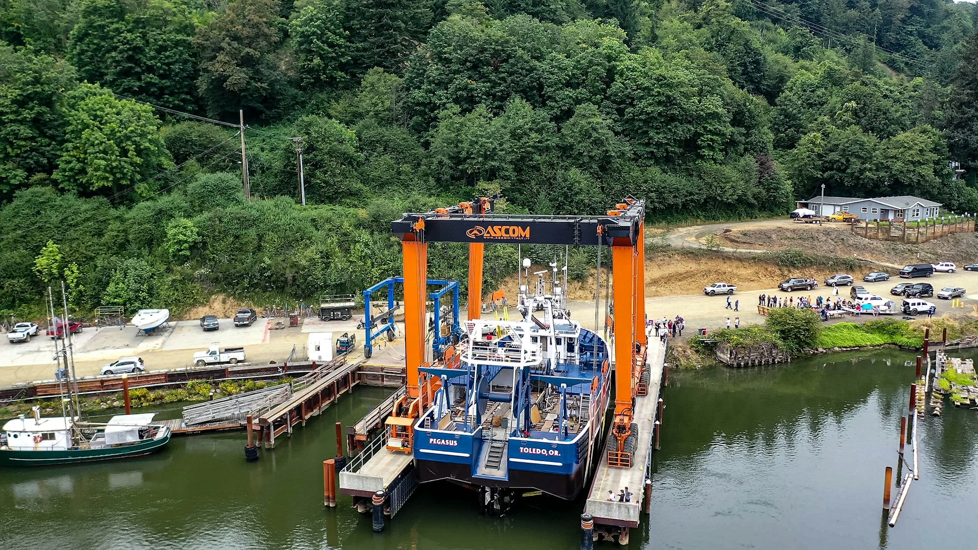 A large blue ship, the "PEGASUS," docked at a pier with an orange crane structure overhead; surrounded by trees, cars, and spectators.