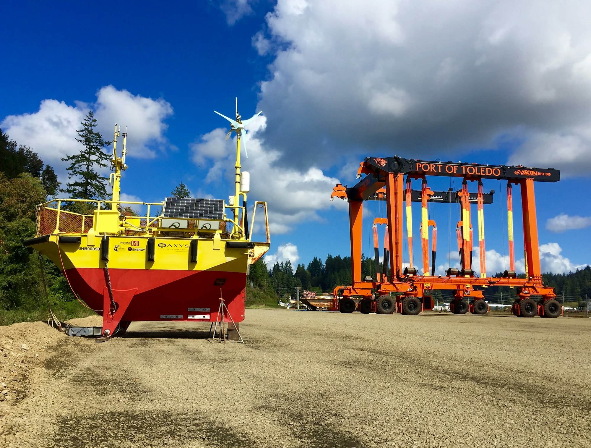 A yellow boat with solar panels and a wind turbine near an orange Port of Toledo gantry on a clear day.