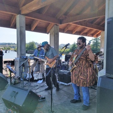 A band performing outdoors under a pavilion with guitars and microphones.