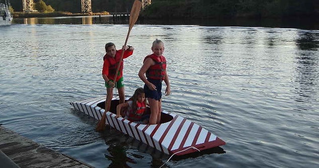 Three kids in life jackets on a handmade boat near a dock.