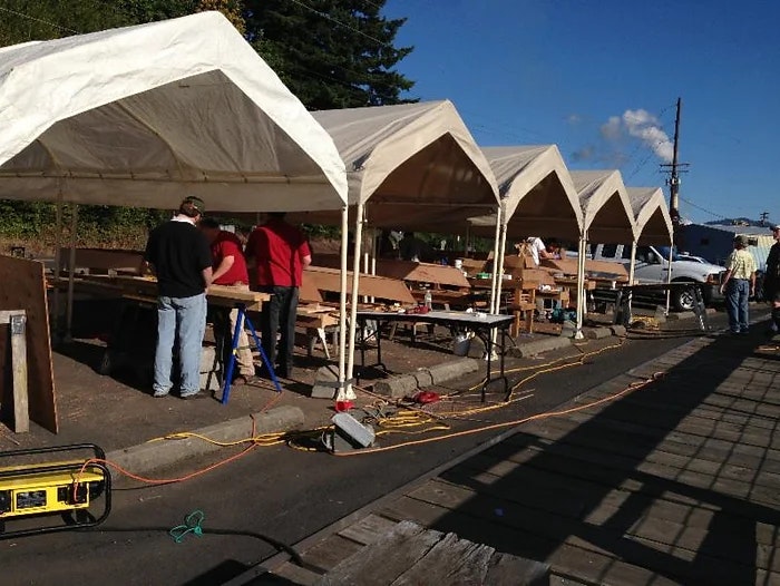 Outdoor workshop with people, tents, tables, tools, and construction material.