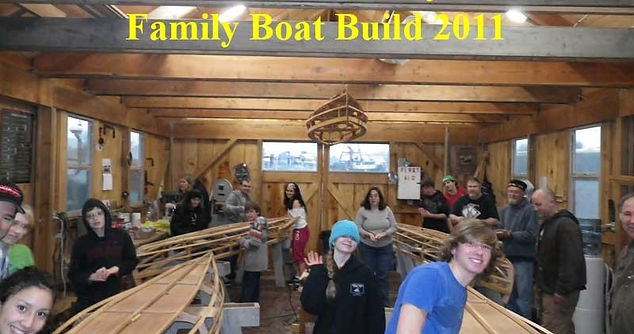 A group of people standing around wooden boat frames, with a banner reading "Family Boat Build 2011."