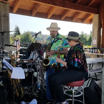 Two musicians playing saxophone and flute outdoors with a drum set in the background.