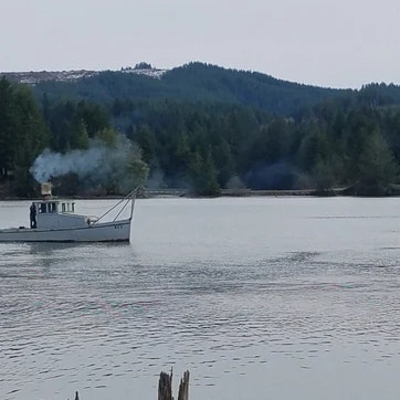 A boat on a calm lake with smoke coming from its chimney against a backdrop of forested hills.