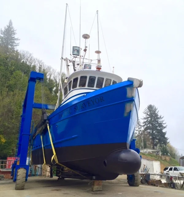 A blue fishing boat named "Pacific Surveyor" on a dry dock with trees and sky in the background.