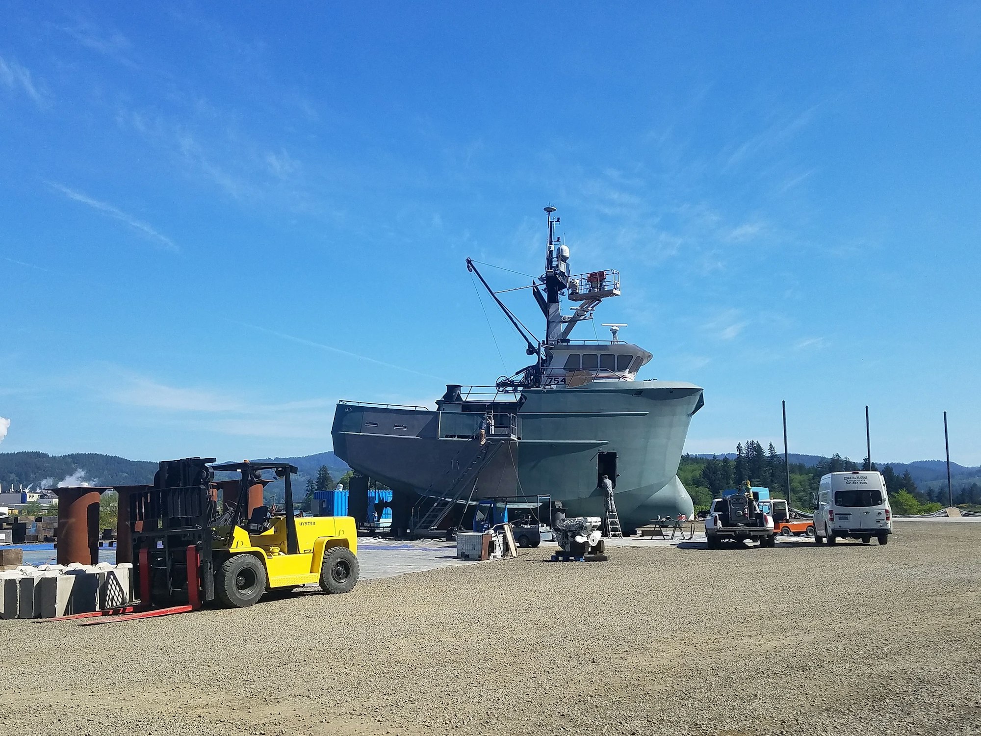 A ship under construction or repair at a dry dock facility, with forklifts and materials around, under a clear blue sky.