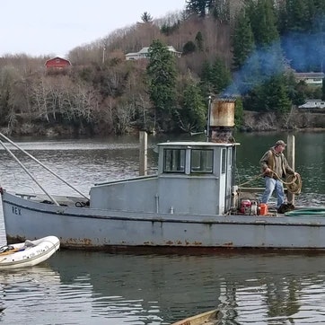 A person on a docked boat with a smoking chimney, near trees and a hill.