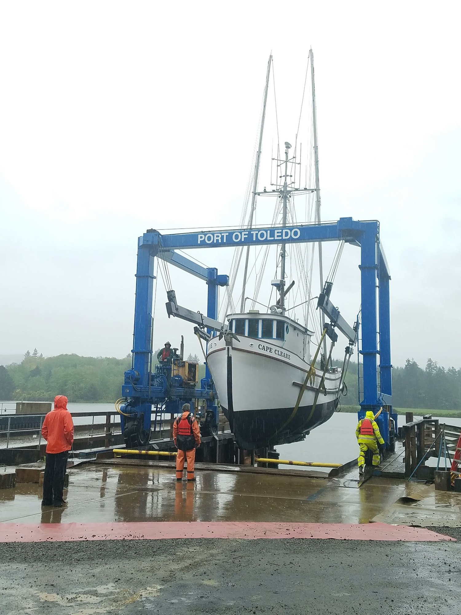 A boat named "CAPE CLEAR" at the Port of Toledo, being hoisted by a blue crane, with three people in rain gear observing.