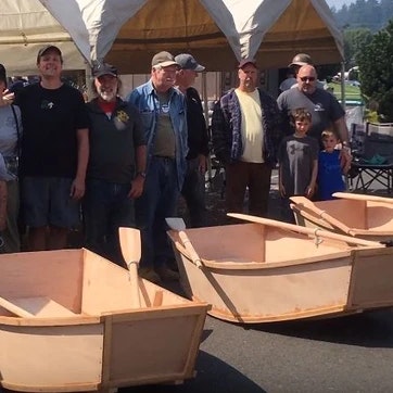 Group of people standing behind handmade wooden boats, likely at an outdoor event or exhibition.