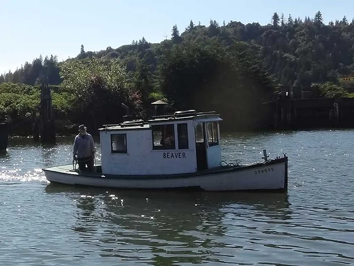 A person on a small boat named "BEAVER" on a river, with trees and a clear sky in the background.