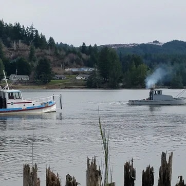 A boat and a barge on a river with smoke rising from the barge, surrounded by trees and broken pilings in the foreground.