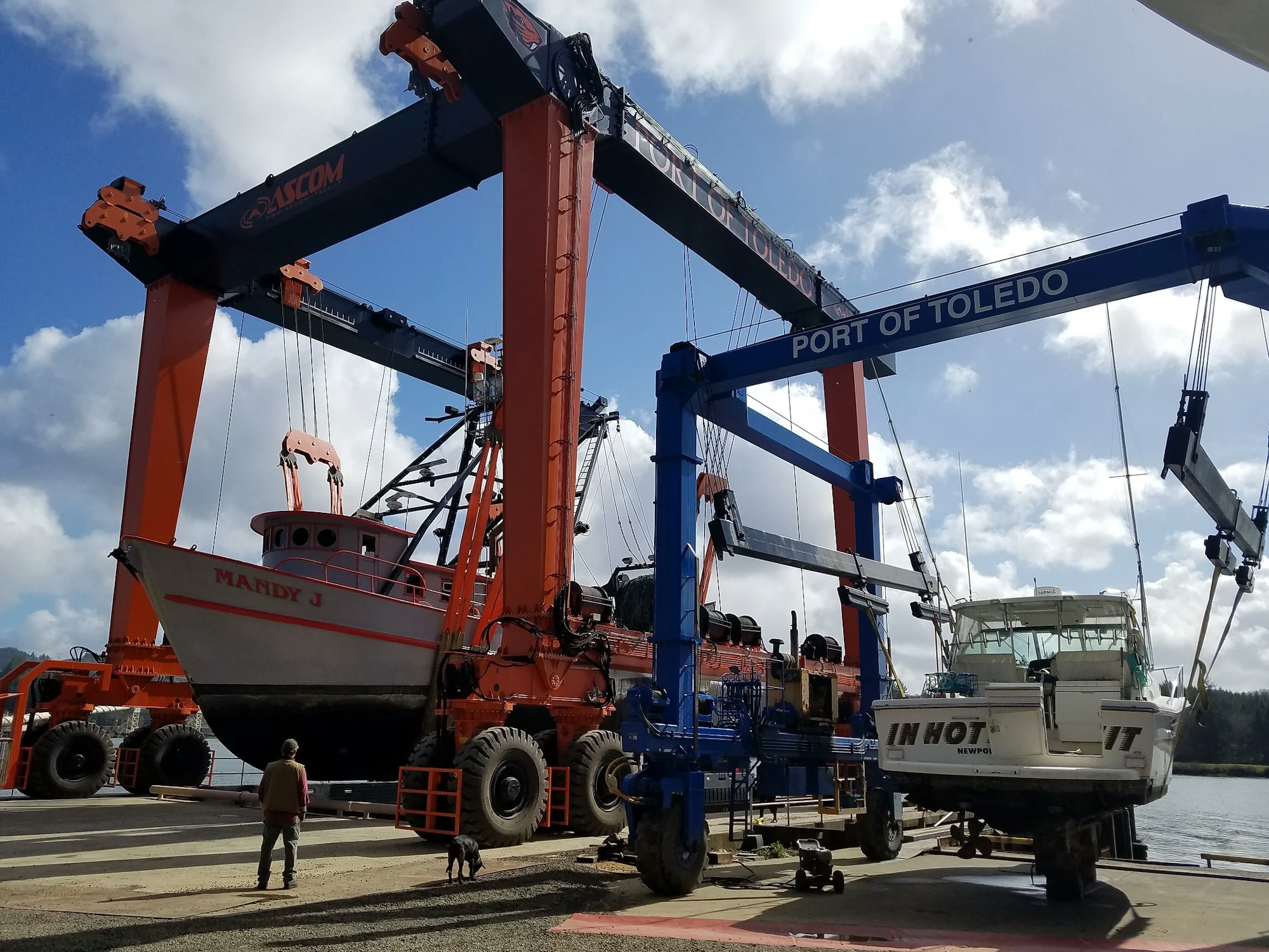 A large mobile boat hoist lifting a boat, with a person and a dog watching, at Port of Toledo.