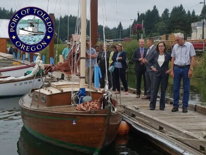 A group of people by a wooden sailboat at a dock, with a "Port of Toledo, Oregon" logo overlay.