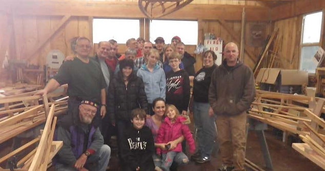 A group of people smiling in a workshop with wooden materials in the background.