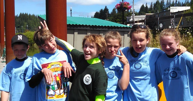 Group of kids posing with playful gestures, wearing matching blue shirts, outdoors on a sunny day.
