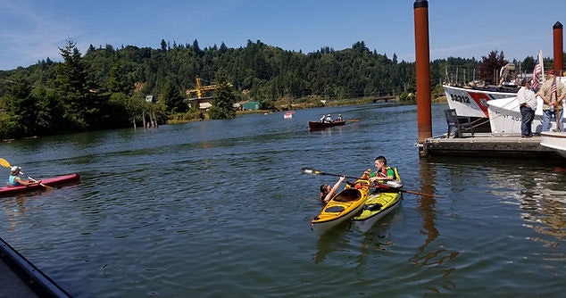People kayaking on a river, a dock on the right, greenery in the background, sunny day.