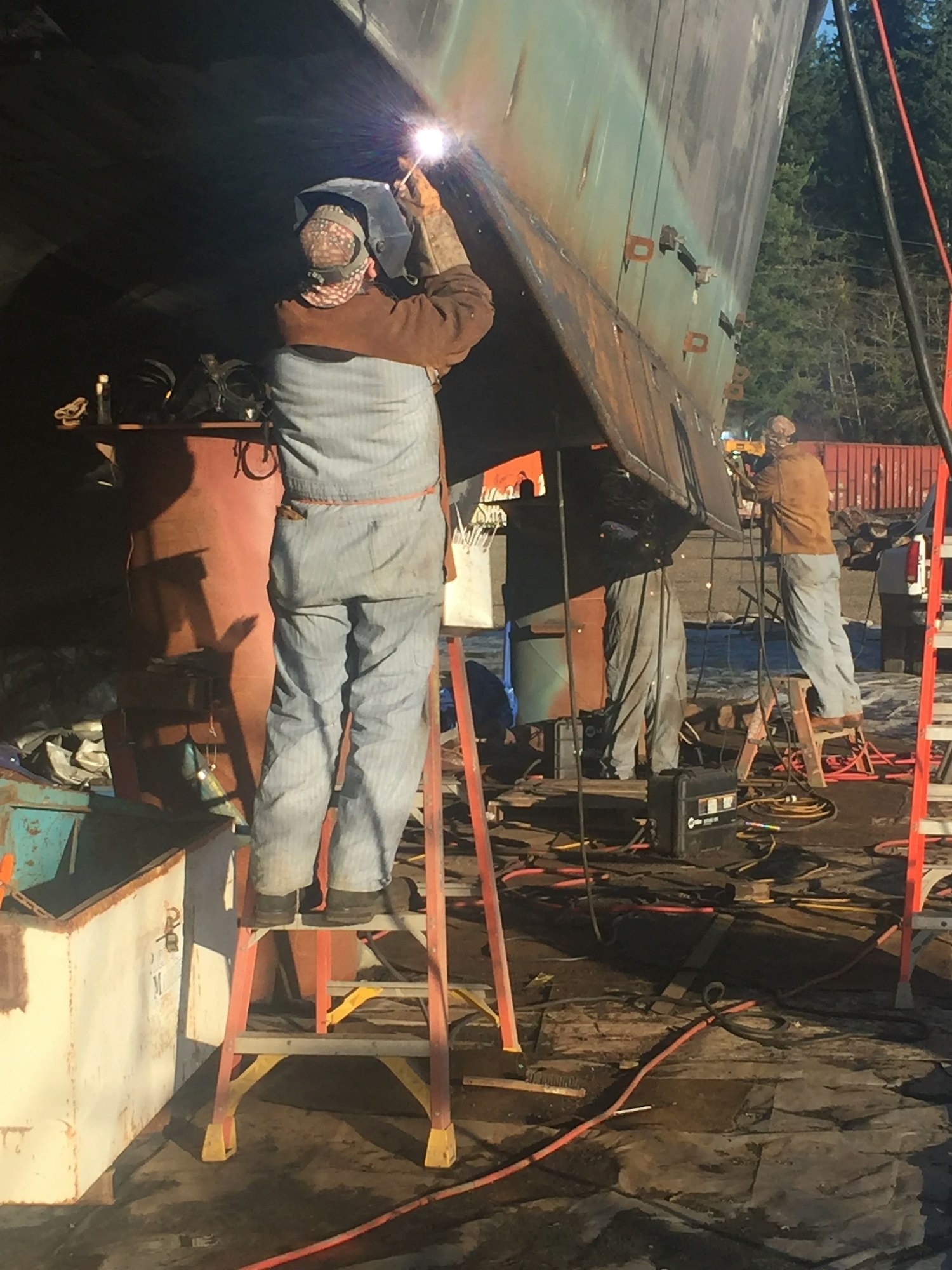 Workers welding on a ship hull, equipment scattered around, outdoors during the day.