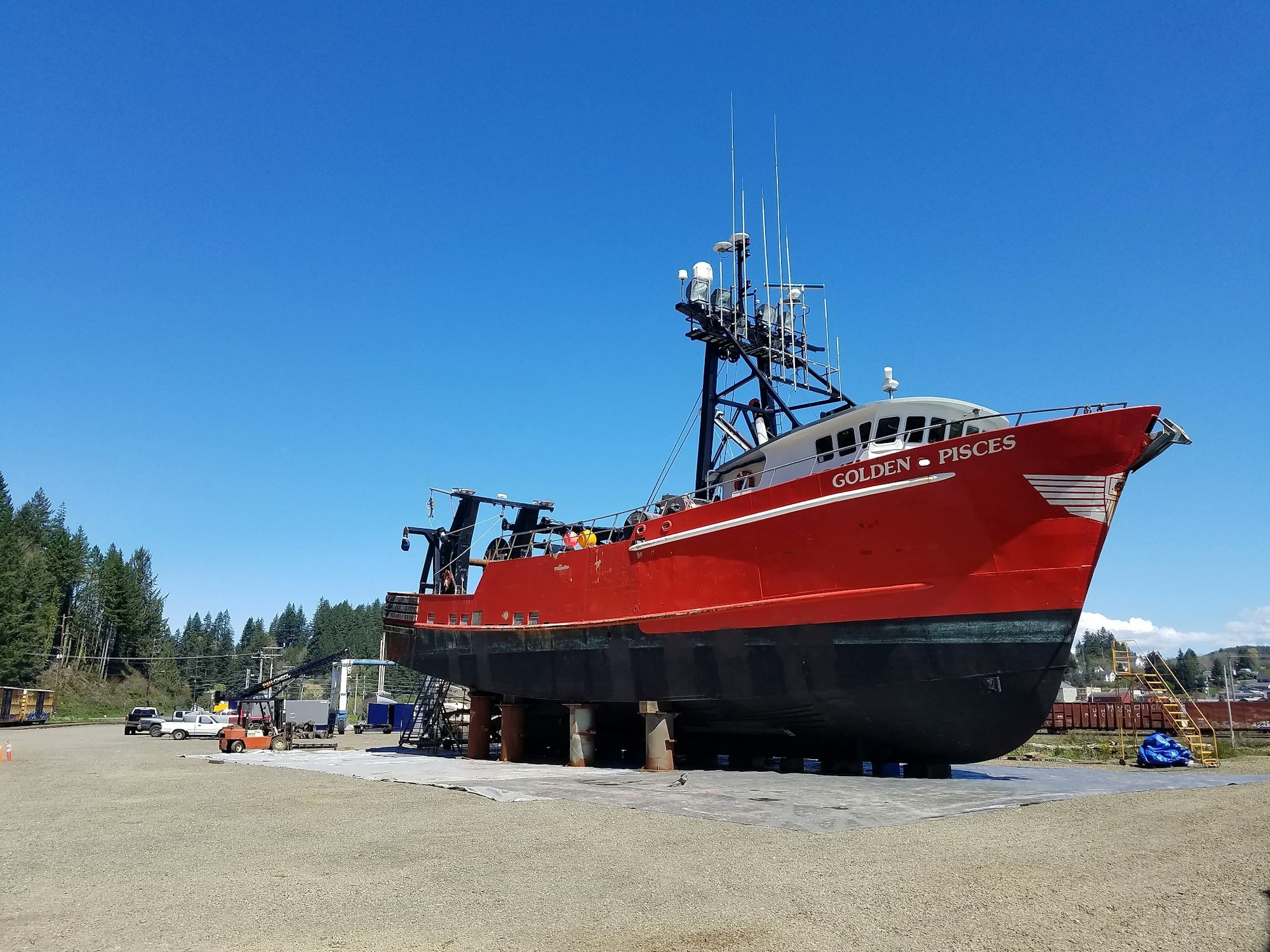 Red fishing vessel "Golden Pisces" on dry land for repairs or maintenance, clear blue sky above.