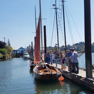 People on a dock beside a sailboat with a red sail, clear skies, and calm water.