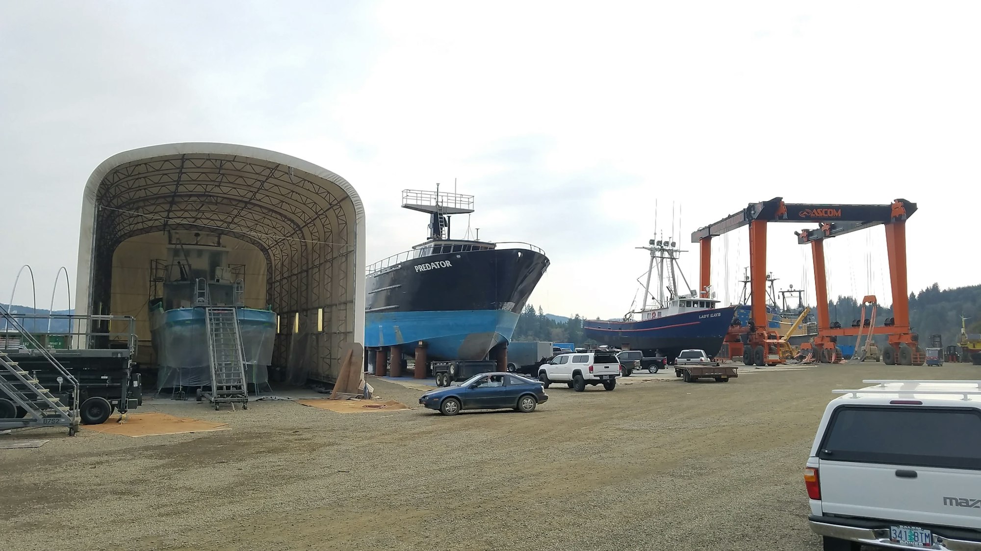 A shipyard with boats in drydock, a large overhead crane system, vehicles, and a covered structure.