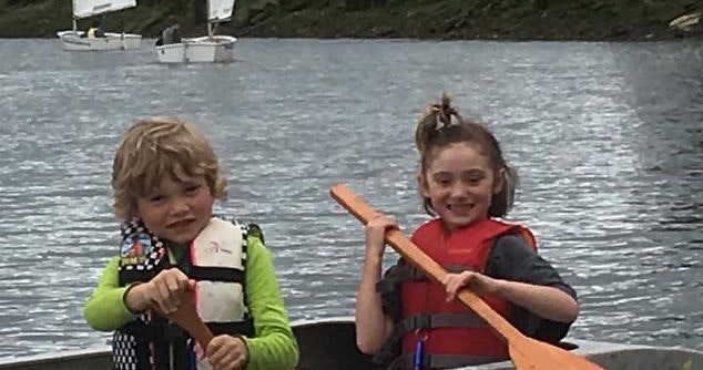 Two children in life vests holding paddles on water with boats in the background.