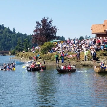 A crowd watches as quirky homemade boats race in a water event on a sunny day.
