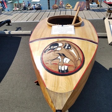 A wooden boat with indigenous-style artwork on the dock, paddle inside, under clear skies. #BoatArt #WoodenCraftsmanship