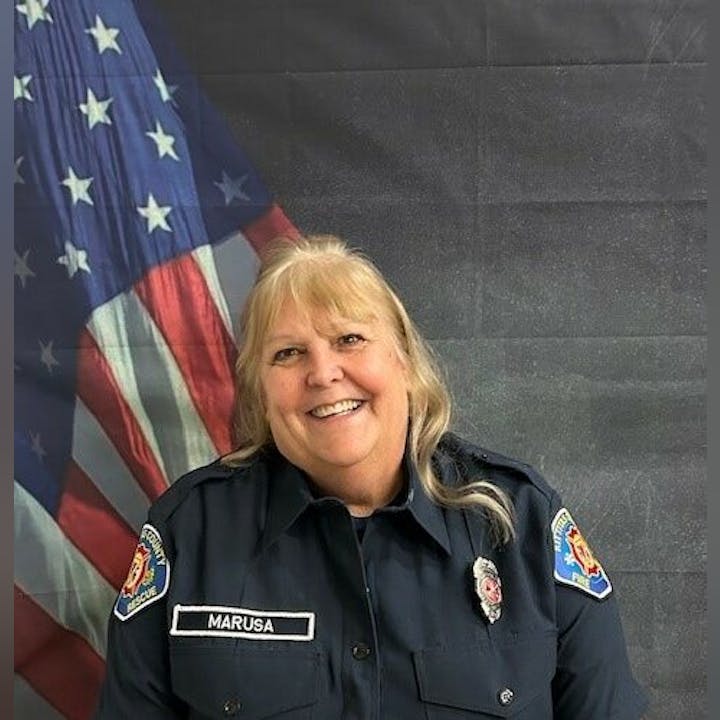 A person in a uniform with name patch "Marusa" stands in front of an American flag backdrop.