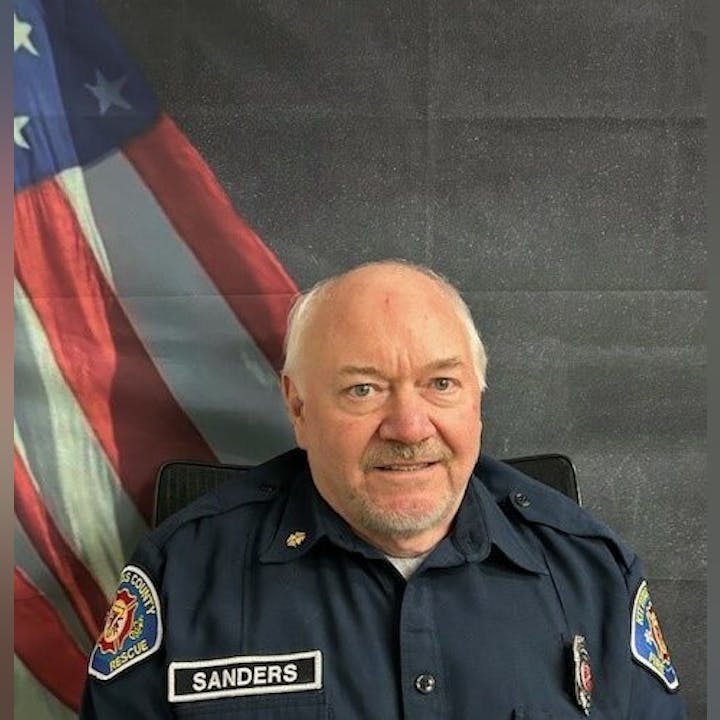 A man in a uniform with "Sanders" on a name patch sits in front of an American flag backdrop.