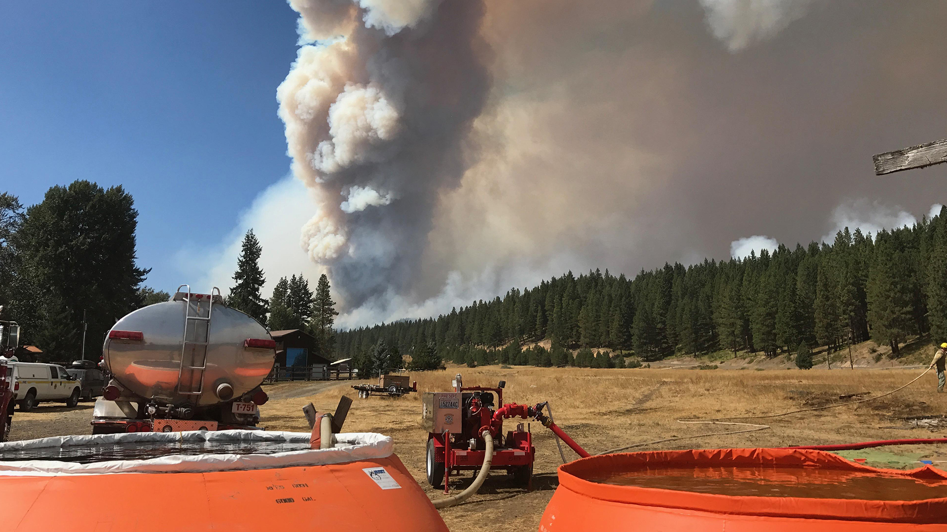 Smoke billows over a forest; firefighting equipment and tanks are visible in the foreground.