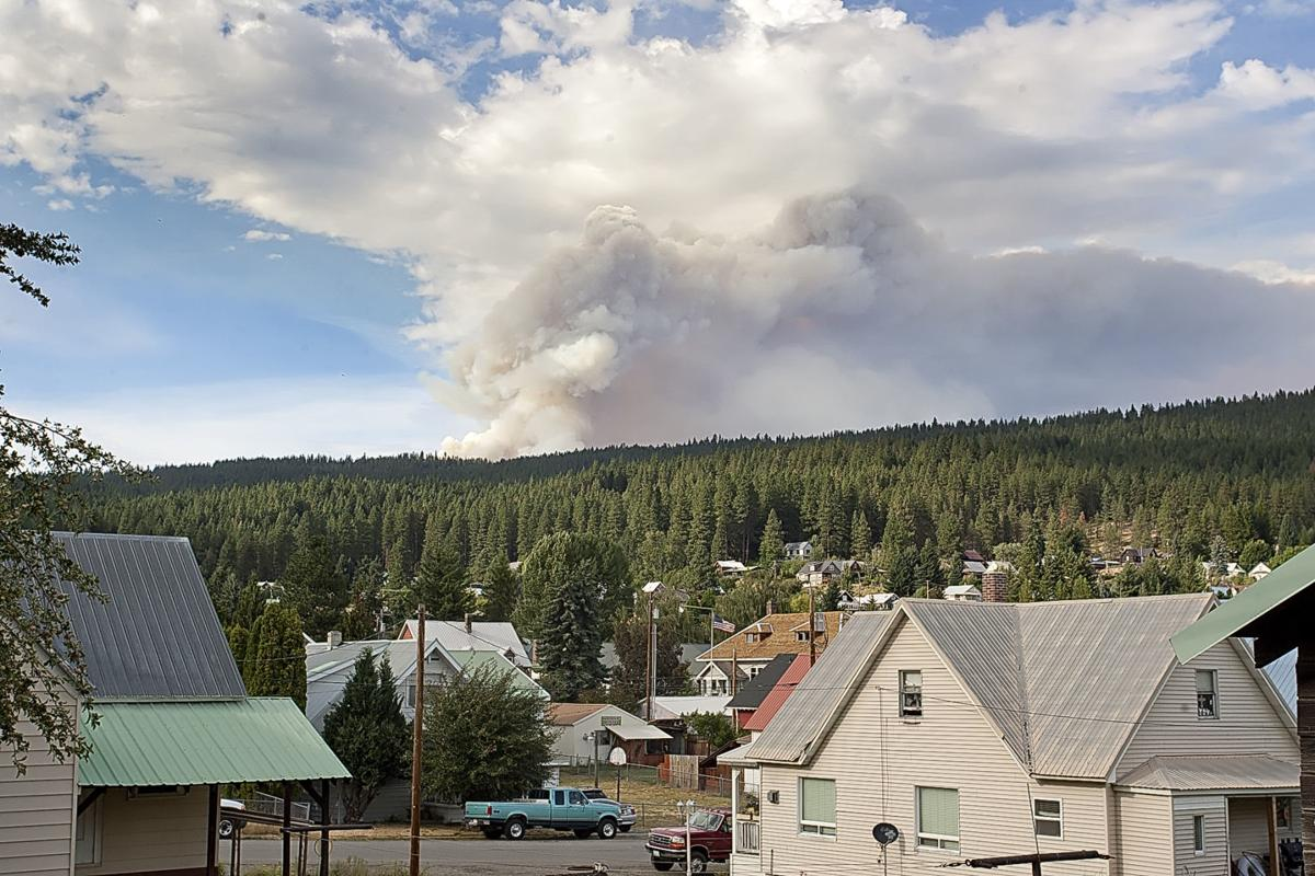 A wildfire is visible behind houses in a forested area, with smoke rising into the sky.