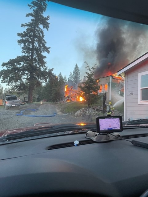 A house on fire with heavy smoke, viewed from inside a vehicle, parked near trees and other buildings.
