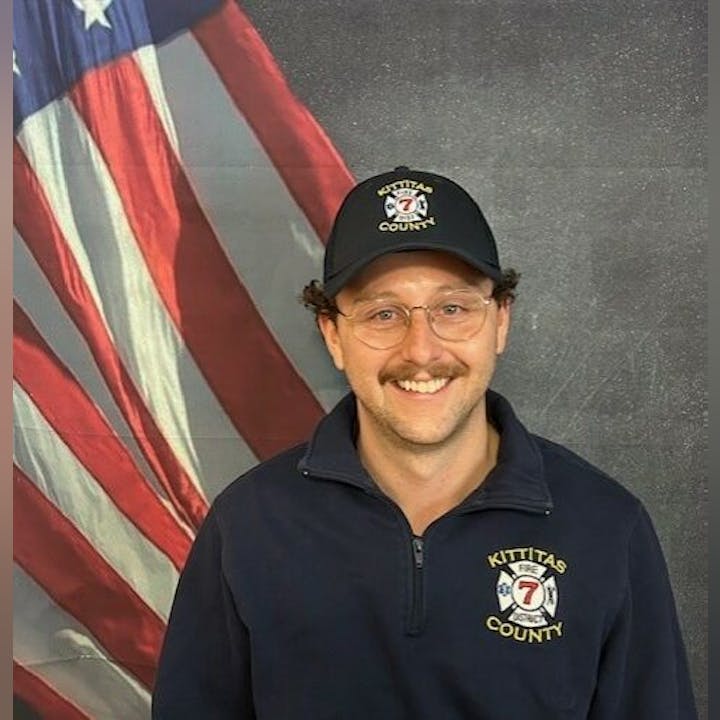Person smiling in front of a U.S. flag, wearing a Kittitas County Fire District hat and jacket.