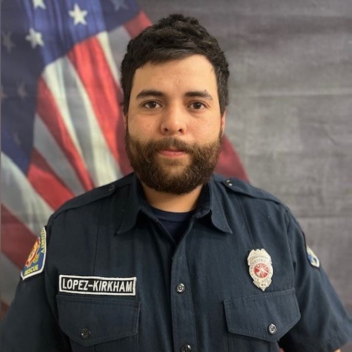 A person in a uniform with a badge, standing in front of an American flag backdrop.