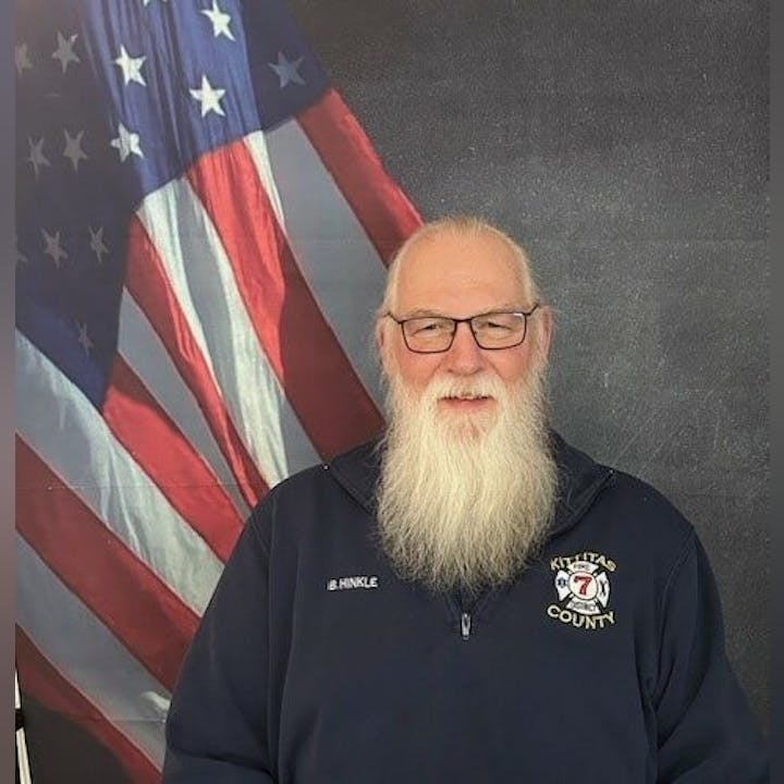 A bearded man in glasses with an American flag backdrop, wearing a "Kittitas County" jacket with a patch and name badge.
