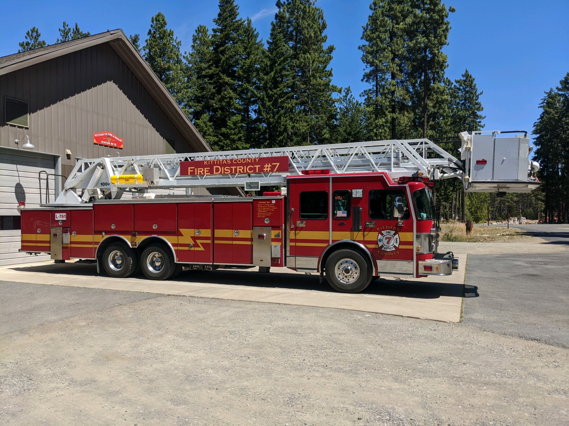 A red fire truck with a ladder outside a fire station, surrounded by trees.