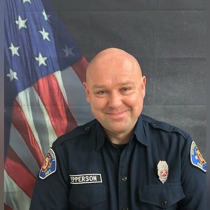 A person in a uniform with badges stands in front of an American flag.