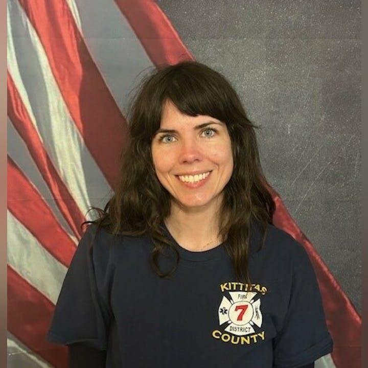 A person smiling in front of an American flag background, wearing a navy T-shirt with a "Kittitas County Fire District 7" emblem.