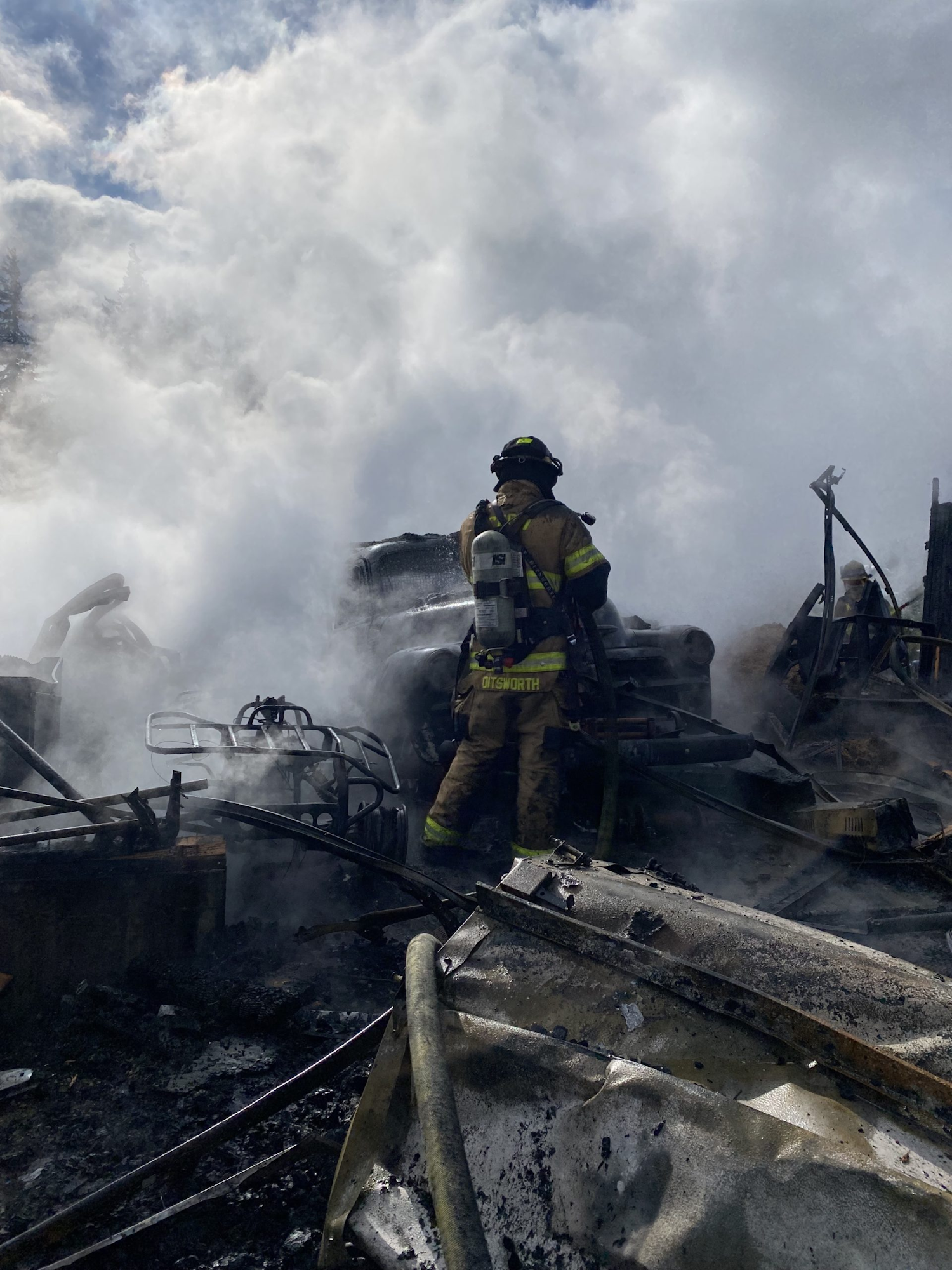 Firefighter in gear stands amidst smoke and debris, working to control a fire.