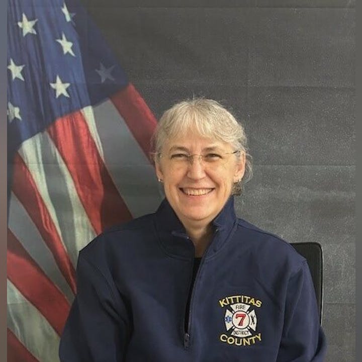 A person in a Kittitas County Fire District jacket, smiling, with a U.S. flag in the background.