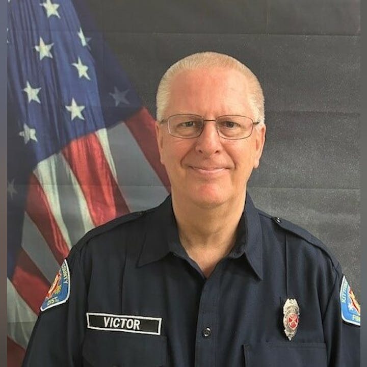 A man in a uniform with a name tag "Victor" stands in front of an American flag.