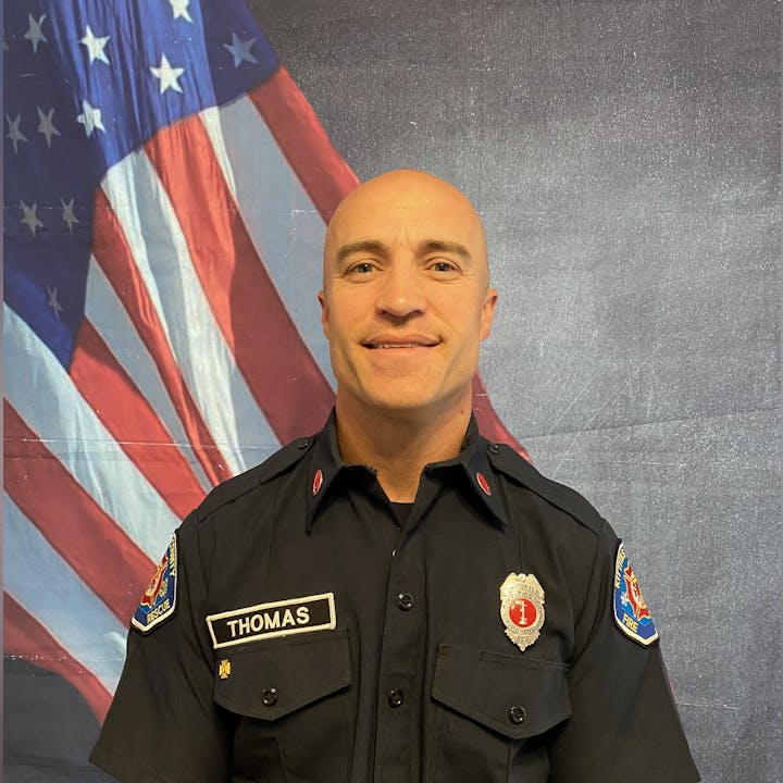 A person in a fire department uniform with "Thomas" on the name tag, standing in front of an American flag background.