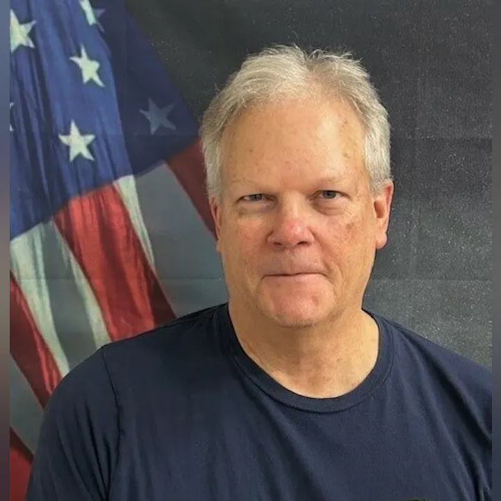 A man in a dark shirt stands in front of an American flag backdrop.