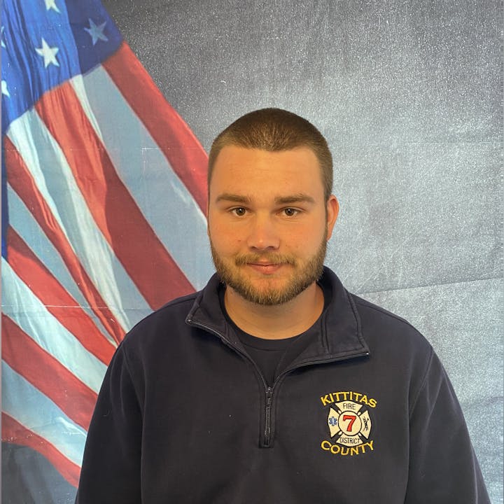 A person in a navy uniform with "Kittitas County Fire District 7" emblem stands before an American flag backdrop.