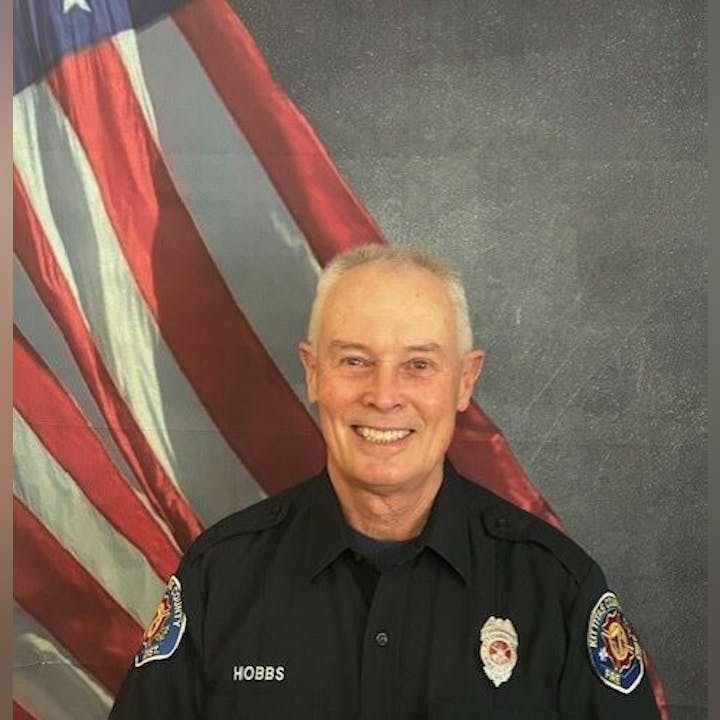 A smiling man in uniform with "HOBBS" on his shirt stands in front of an American flag backdrop.