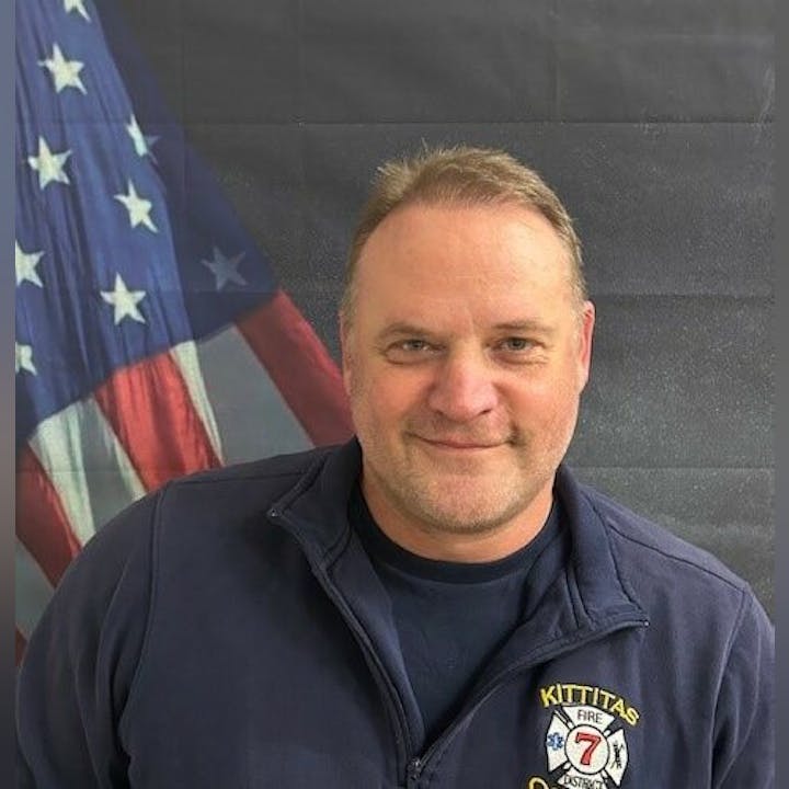 A person in a navy jacket with "Kittitas Fire & Rescue" emblem stands in front of a U.S. flag background.