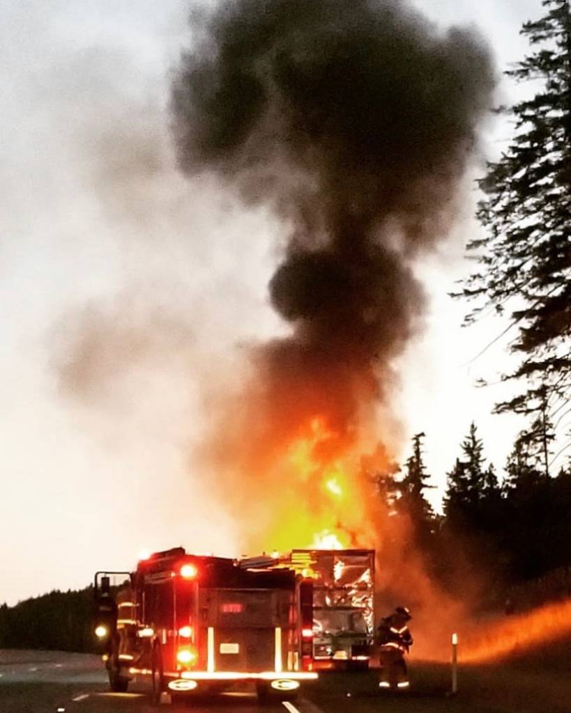 Firetruck and firefighter at a truck fire with large flames and smoke on the side of a road surrounded by trees.