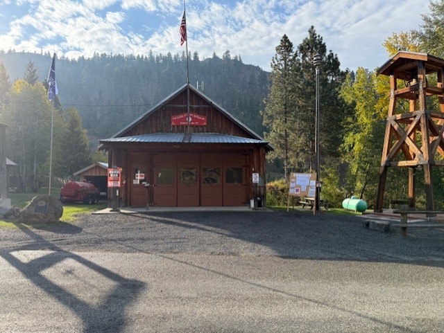 A wooden fire station with flags, surrounded by trees and mountains under a blue sky.